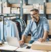 Smiling man in glasses working on a laptop in a clothing webshop, surrounded by boxes and hanging garments