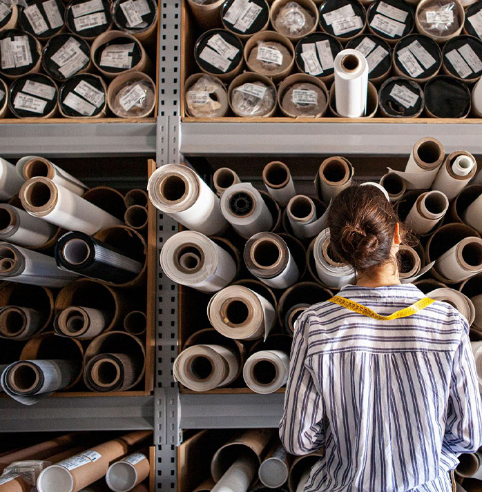 Person with a measuring tape around their neck, standing in front of shelves filled with rolled fabrics