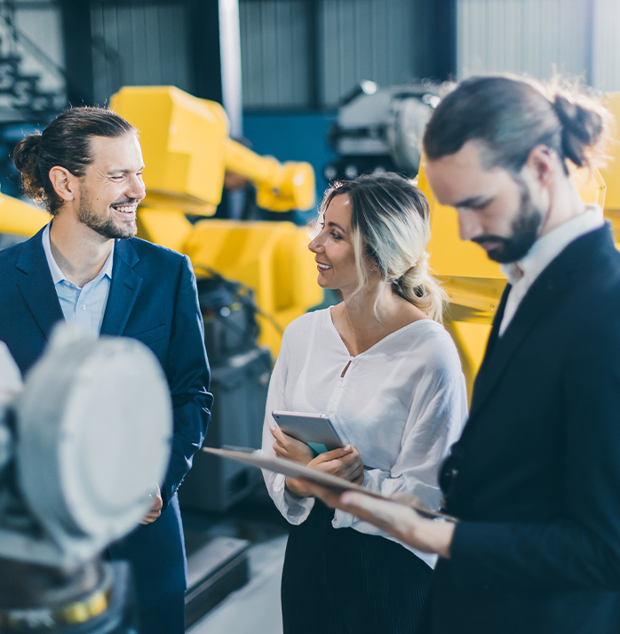 Three colleagues in a discussion, standing in front of yellow industrial machinery, with one person holding a tablet