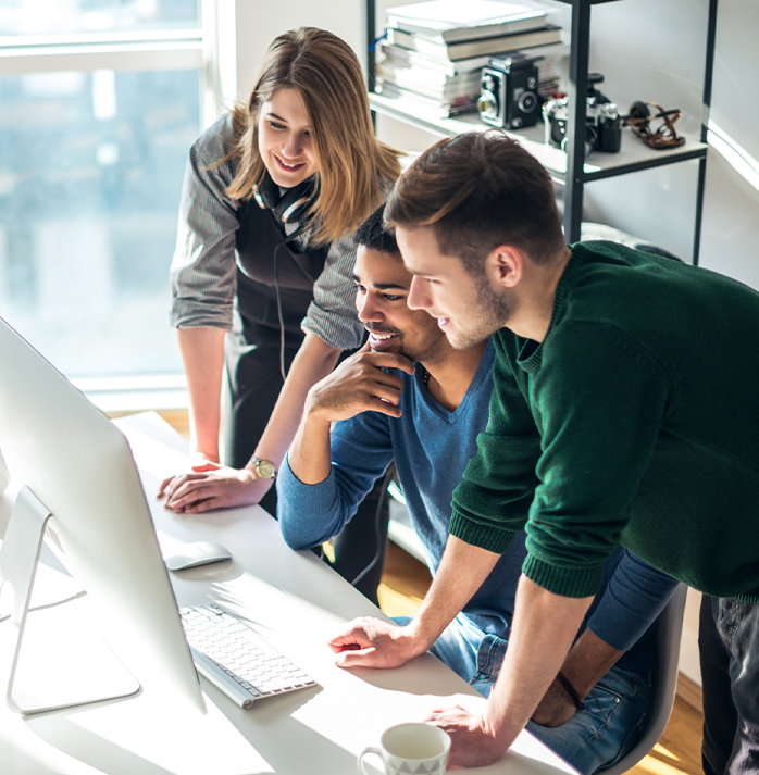 Three colleagues smiling and collaborating around a desktop computer in a bright, modern office